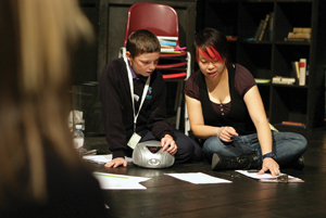 A teacher and a student in uniform sit on the floor and look at papers spread out in front of them